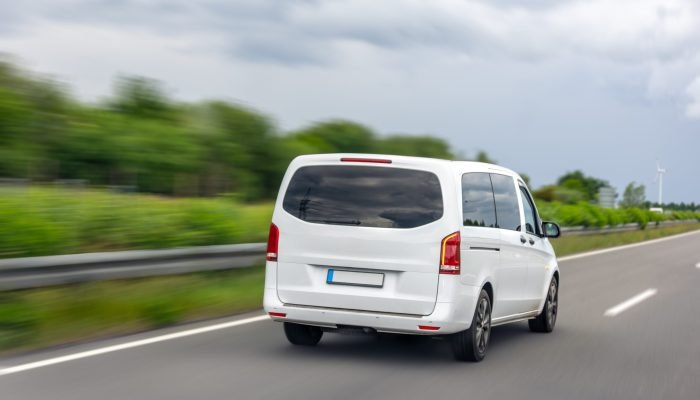 A white minivan driving on an asphalt highway with blurred background of trees and cloudy sky. Rear view capture.